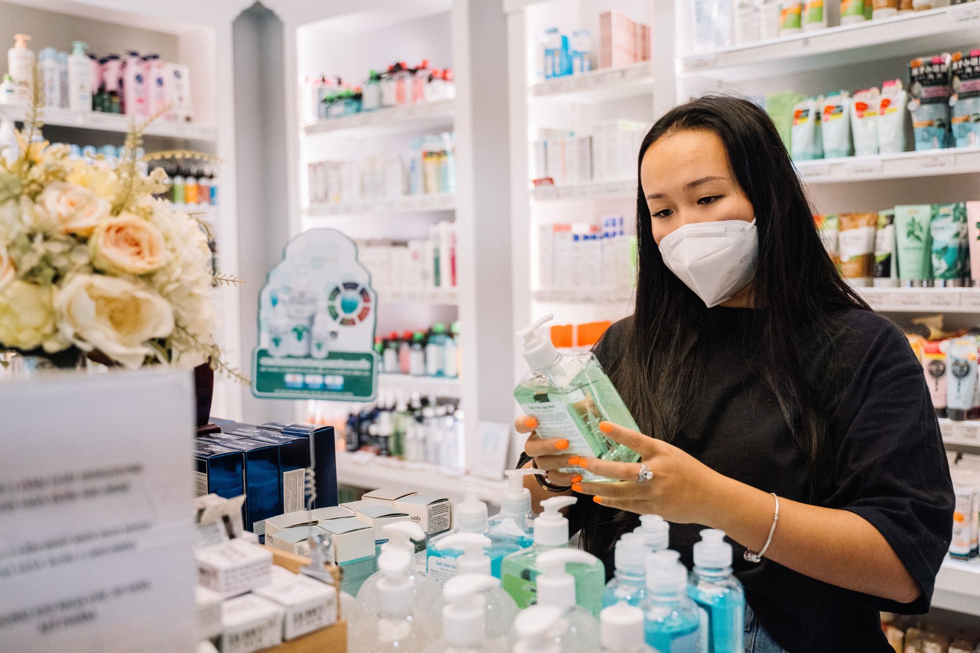 Femme portant un masque, examinant un produit désinfectant dans une officine moderne, entourée d'étagères remplies de produits de santé et de soins.