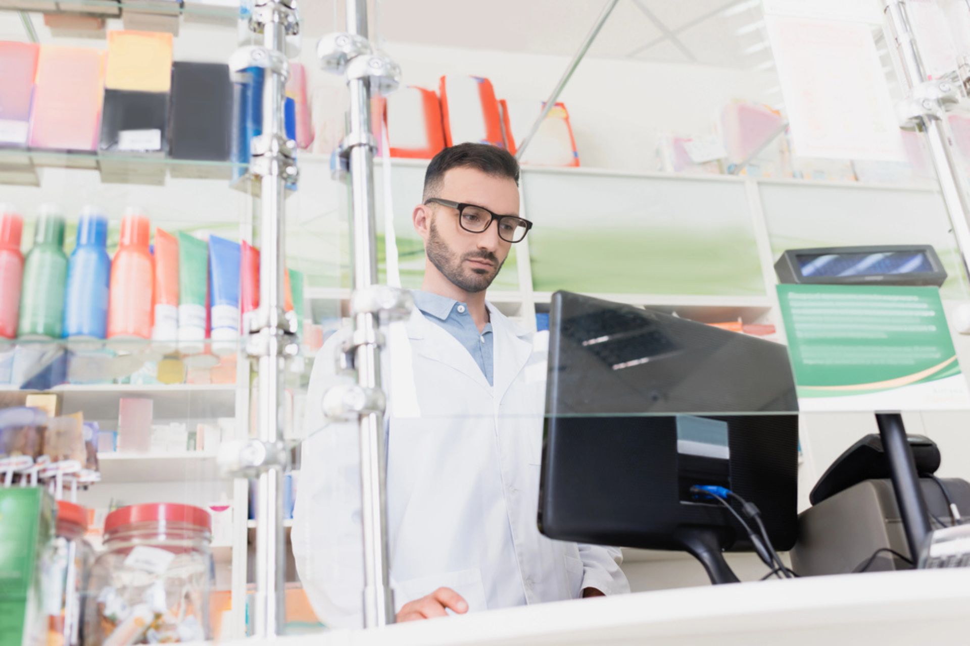 Pharmacien consultant un logiciel de gestion sur un ordinateur derrière le comptoir d’une officine, illustrant la digitalisation des audits internes en pharmacie.
